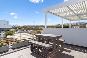 Terraza en la azotea con mesa y banco de madera, zona cubierta y vista a un barrio residencial.