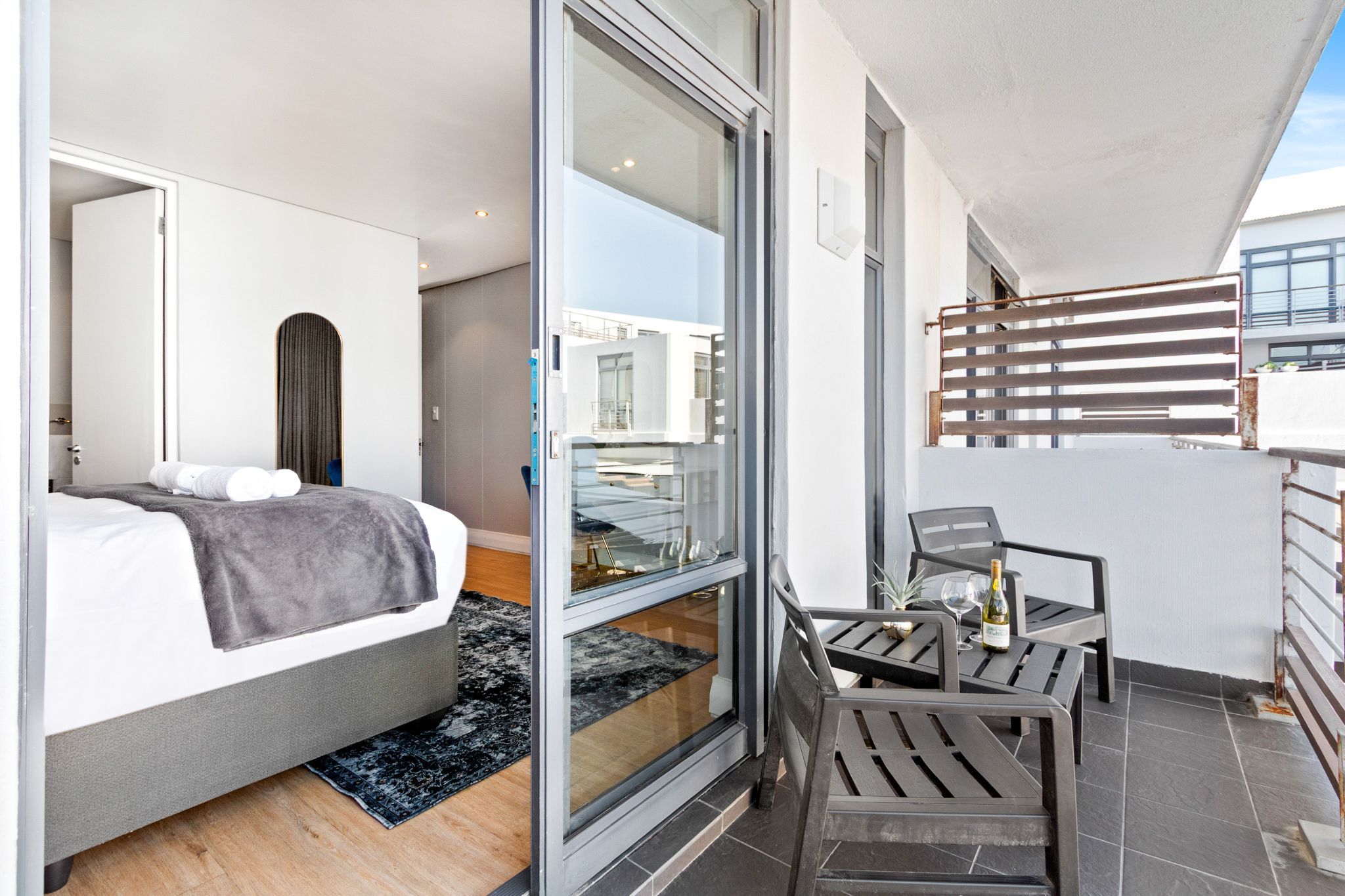 A modern bedroom with a bed, a glass door leading to a balcony, and a small table with a bottle and glasses.
