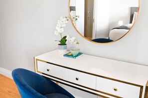 A white dresser with gold trim, a blue chair, a potted orchid, and a round mirror with a gold frame. - Eden on the Bay 190 (Cape Town)