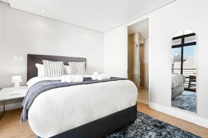 A modern hotel room with a large bed, a bedside table with a lamp, a mirror, and a view of the outside. - Eden on the Bay 190 (Cape Town)