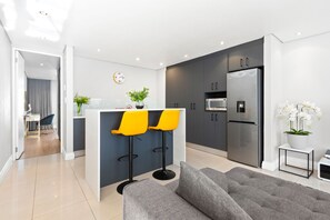A modern kitchen with a central island, two yellow bar stools, a grey sofa, and a small side table with a plant. - Eden on the Bay 190 (Cape Town)