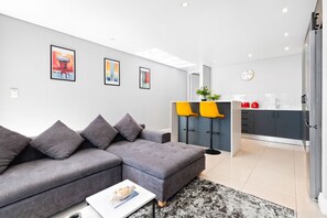A modern living room with a grey sectional sofa, a coffee table, and a black and white rug. There are framed artworks on the wall and a kitchen area with grey cabinets and yellow bar stools. - Eden on the Bay 190 (Cape Town)