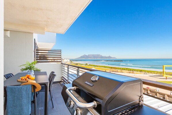 A balcony with a grill, dining table, chairs, and a view of the ocean. - Eden On The Bay 168 (Cape Town)