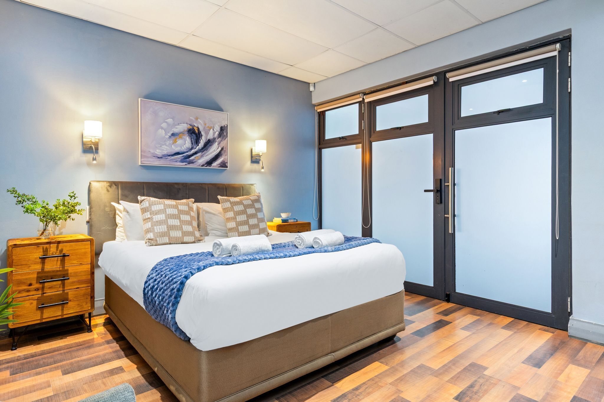 A bedroom with a large bed, a wooden dresser, a wall-mounted lamp, and a painting.