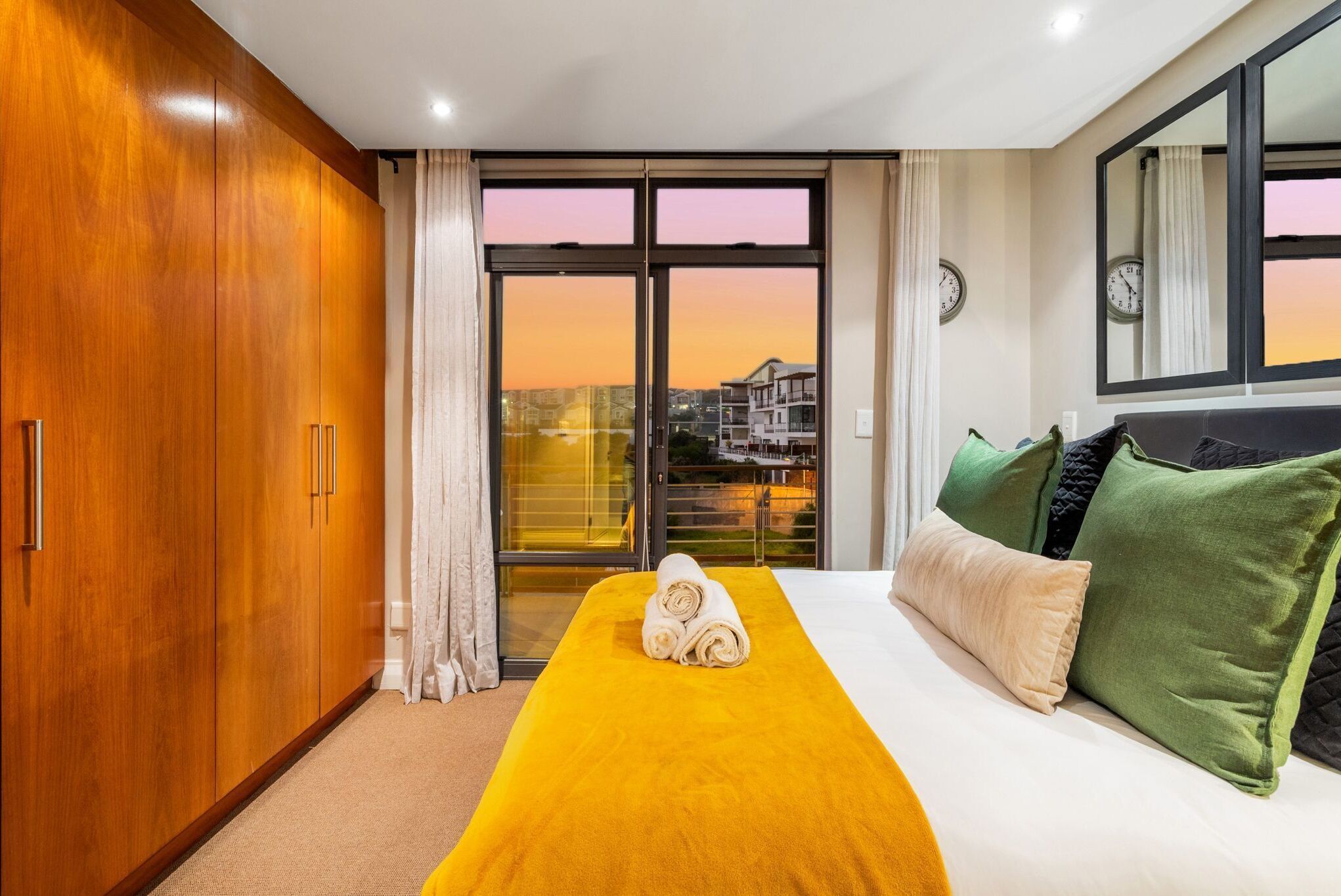 A hotel room with a large bed, a wooden wardrobe, a balcony with a city view, and a window with curtains.