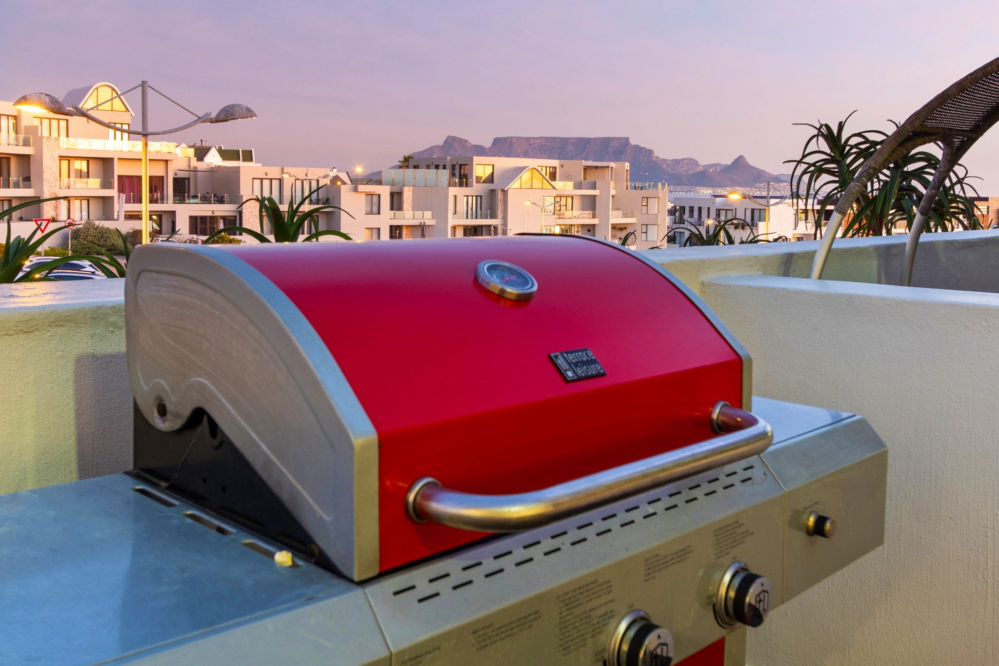 A red barbecue grill with a handle and control knobs, set against a backdrop of modern buildings and a mountain.
