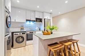 A modern kitchen with built-in appliances, a central island with stools, and a potted plant. - Eden On The Bay 102 (Cape Town)