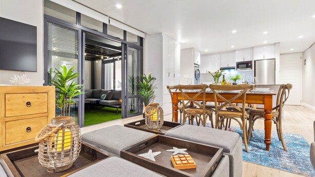 Ein modernes Wohnzimmer mit Essbereich, einer Küche im Hintergrund und einer großen Schiebeglastür, die auf einen mit Pflanzen bepflanzten Balkon führt.