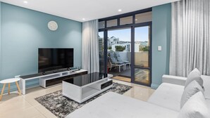 Un salon moderne doté d’un téléviseur à écran plat, d’un canapé blanc et d’une porte vitrée donnant sur un balcon aménagé avec du mobilier d’extérieur.