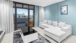Un salon moderne avec un canapé blanc, une table basse en verre et une vue sur un quartier résidentiel, visible à travers de grandes portes coulissantes.