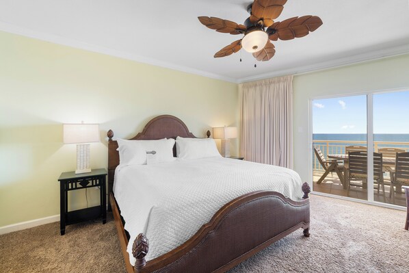 A bedroom with a large bed, a nightstand, a ceiling fan, and a view of the ocean through a sliding glass door.