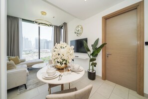 A modern living room with a dining area, a large window with curtains, a flat-screen TV, and a wooden door. - PVH - Tranquil 1BR - Sobha Waves (Dubai)