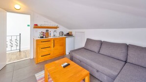 Ett modernt vardagsrum med en grå soffa, ett träbord och en köksdel med träskåp och ett vitt kylskåp.