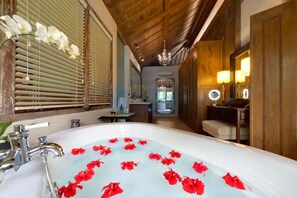 A bathroom with a bathtub filled with water and red flowers, wooden walls, a chandelier, and a mirror with lights. - Kudus Boutique Hotel (Tibubeneng)