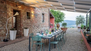 An outdoor dining area with a stone wall, a long table set for dining, and a view of the landscape.