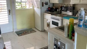 Una cuina compacta amb armaris blancs, un refrigerador, un forn i un rentapeus. Hi ha diversos objectes de cuina i ampolles damunt del taulell.
