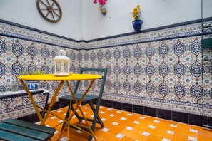 Un angolo con un tavolo giallo e una sedia verde, una parete bianca con piastrelle a motivi blu e arancioni, e un vaso blu con fiori.