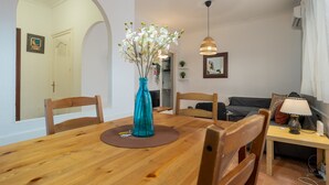 Een eetgedeelte met een houten tafel, stoelen, een vaas met bloemen, een hanglamp en een bank op de achtergrond.