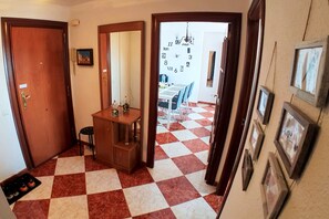 Um corredor com piso xadrez, uma porta de madeira, uma pequena escrivaninha com espelho e uma área de refeições visível através de uma porta aberta.
