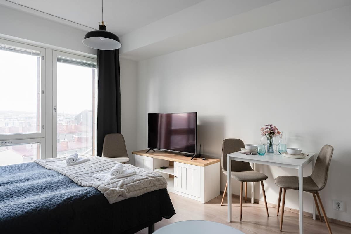 A modern hotel room with a bed, a desk with a TV, a chair, and a dining table with a vase of flowers.