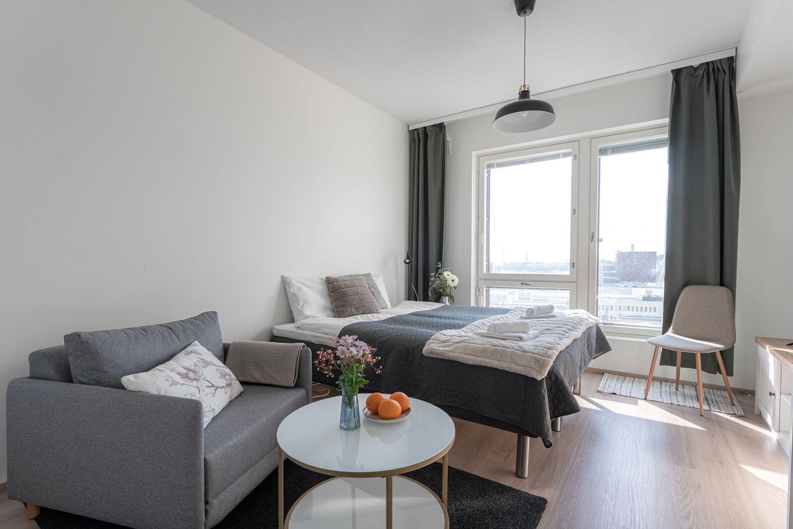 A modern hotel room with a sofa, a bed with a dark duvet, a small round table with a vase and oranges, and a view of the city through large windows.