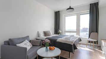 A modern hotel room with a sofa, a bed with a dark duvet, a small round table with a vase and oranges, and a view of the city through large windows.