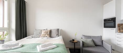 A modern bedroom with a bed, a grey armchair, a small table with a vase, and a built-in oven.