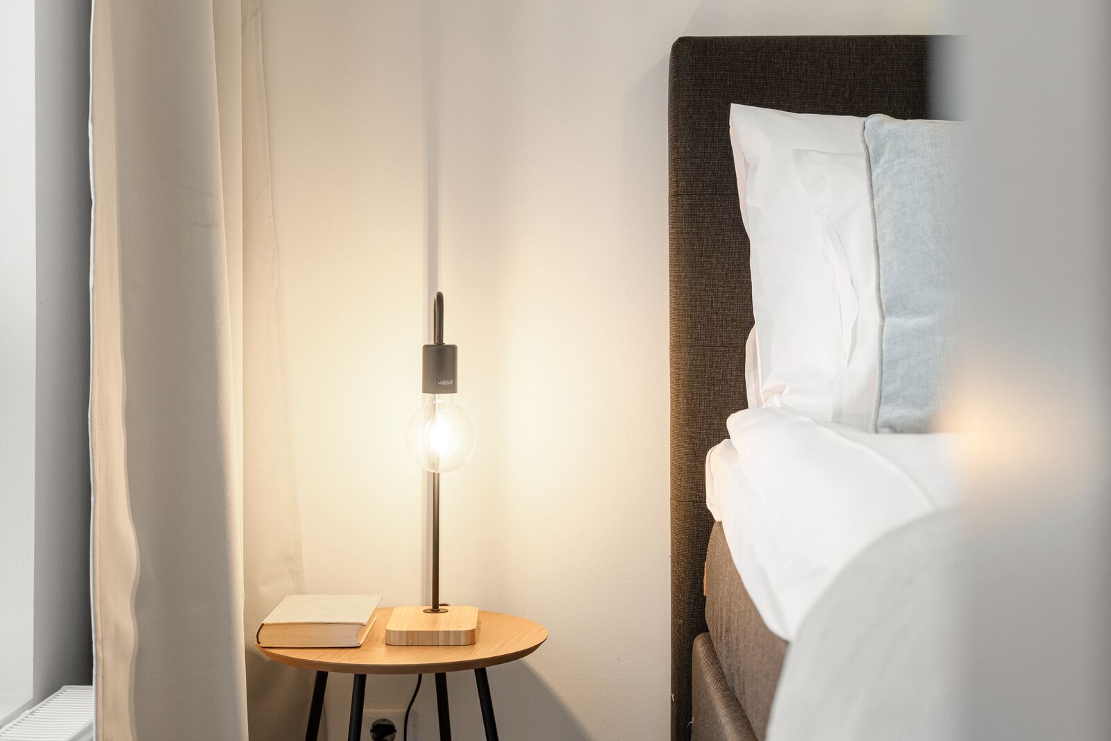 Una cama bien tendida con sábanas blancas, una mesita de noche con un libro y una lámpara, y una lámpara de pared.