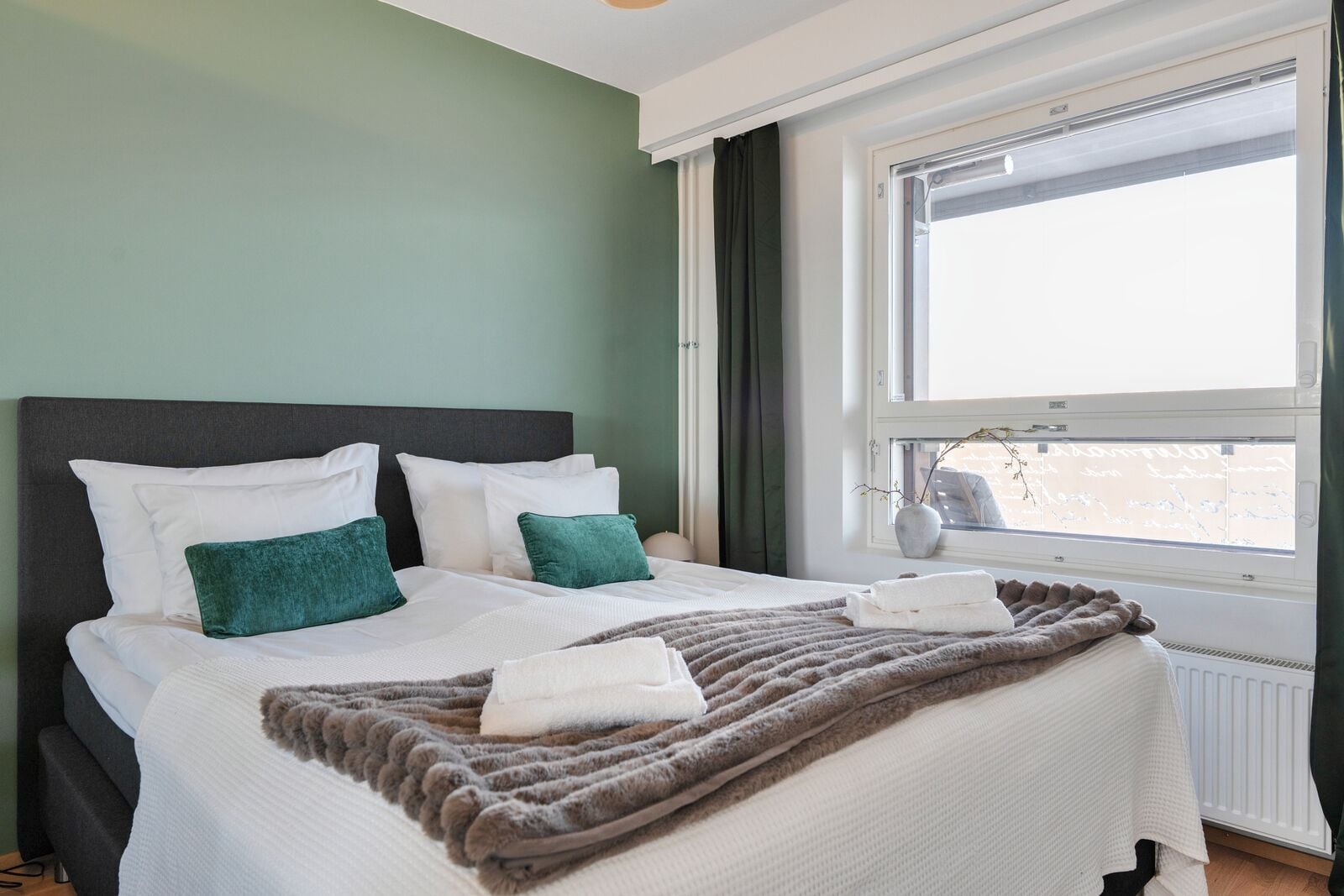 Een netjes opgemaakt bed met witte en groene kussens, een zachte bruine deken en een raam met uitzicht op een besneeerd landschap.