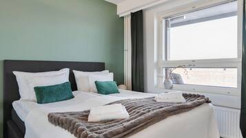 Een netjes opgemaakt bed met witte en groene kussens, een zachte bruine deken en een raam met uitzicht op een besneeerd landschap.