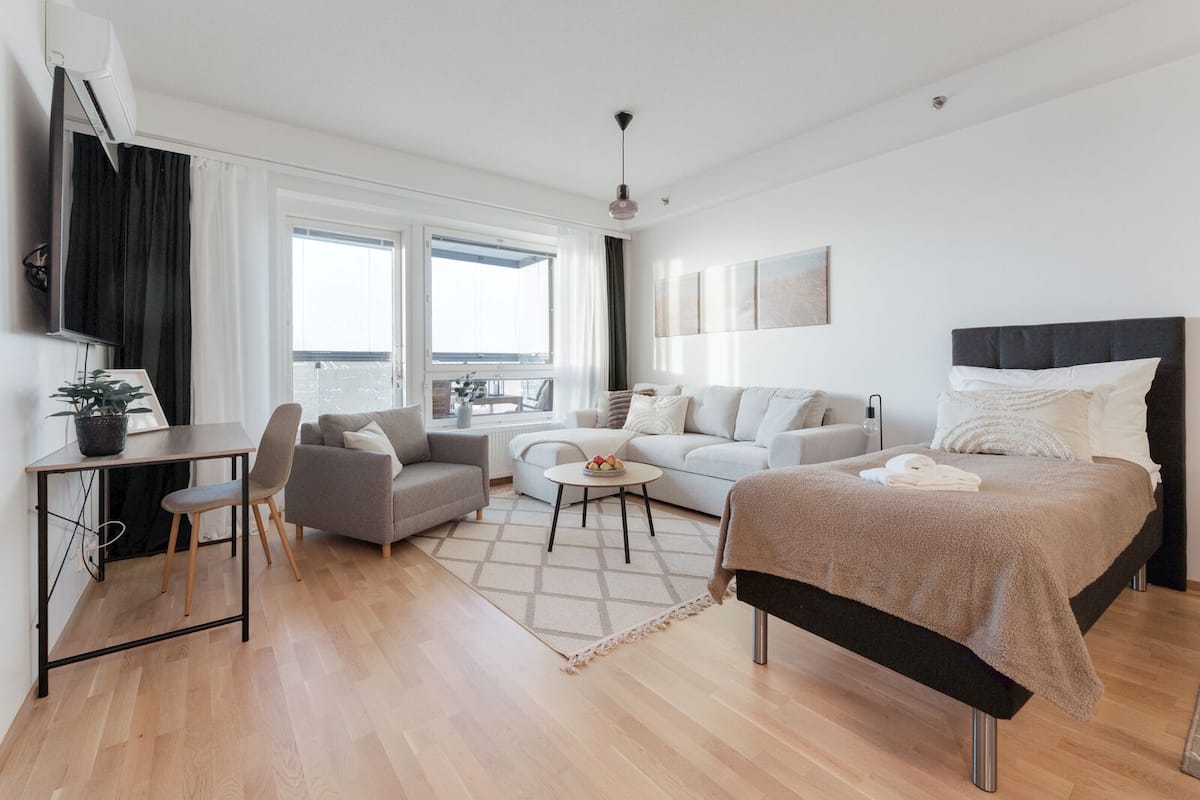 A modern, well-lit living room with a sofa, a dining table with chairs, a bed with a headboard, and a television on the wall.