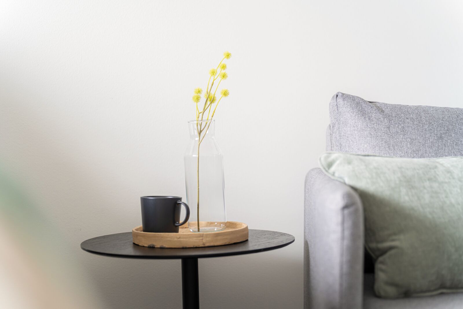 A small round table with a clear glass vase containing yellow flowers, a black mug, and a wooden coaster.