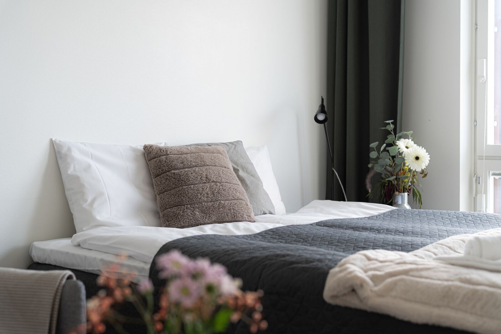 Una cama bien hecha con un edredón gris, una almohada blanca y una almohada marrón texturizada. Hay un florero con flores sobre la mesita de noche.