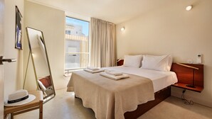 A hotel room with a large bed, a bedside table with a lamp, a mirror, and a view of buildings outside the window. - History and Comfort in the Heart of Vila Real (Vila Real de Santo António)