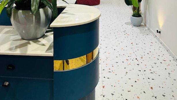 A reception area with a curved blue counter, potted plants, a red sofa, and a hallway with framed artwork and a terrazzo floor.