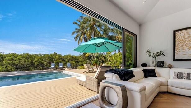 A modern living room with a white sofa, a large window overlooking a pool area with lounge chairs and a green umbrella. - SERENITY ON SUNRISE~Canal Front 5 BR/5.5 Ba New Town Home w/ Pool & Dockage! (Key West)