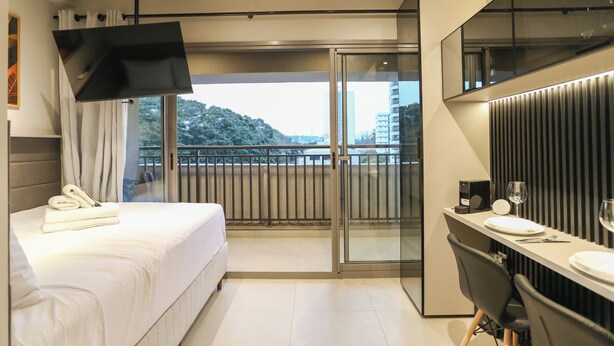 A modern hotel room with a bed, a desk with a chair, a TV, and a balcony with a view of the city. - ON Brooklin (Sao Paulo)