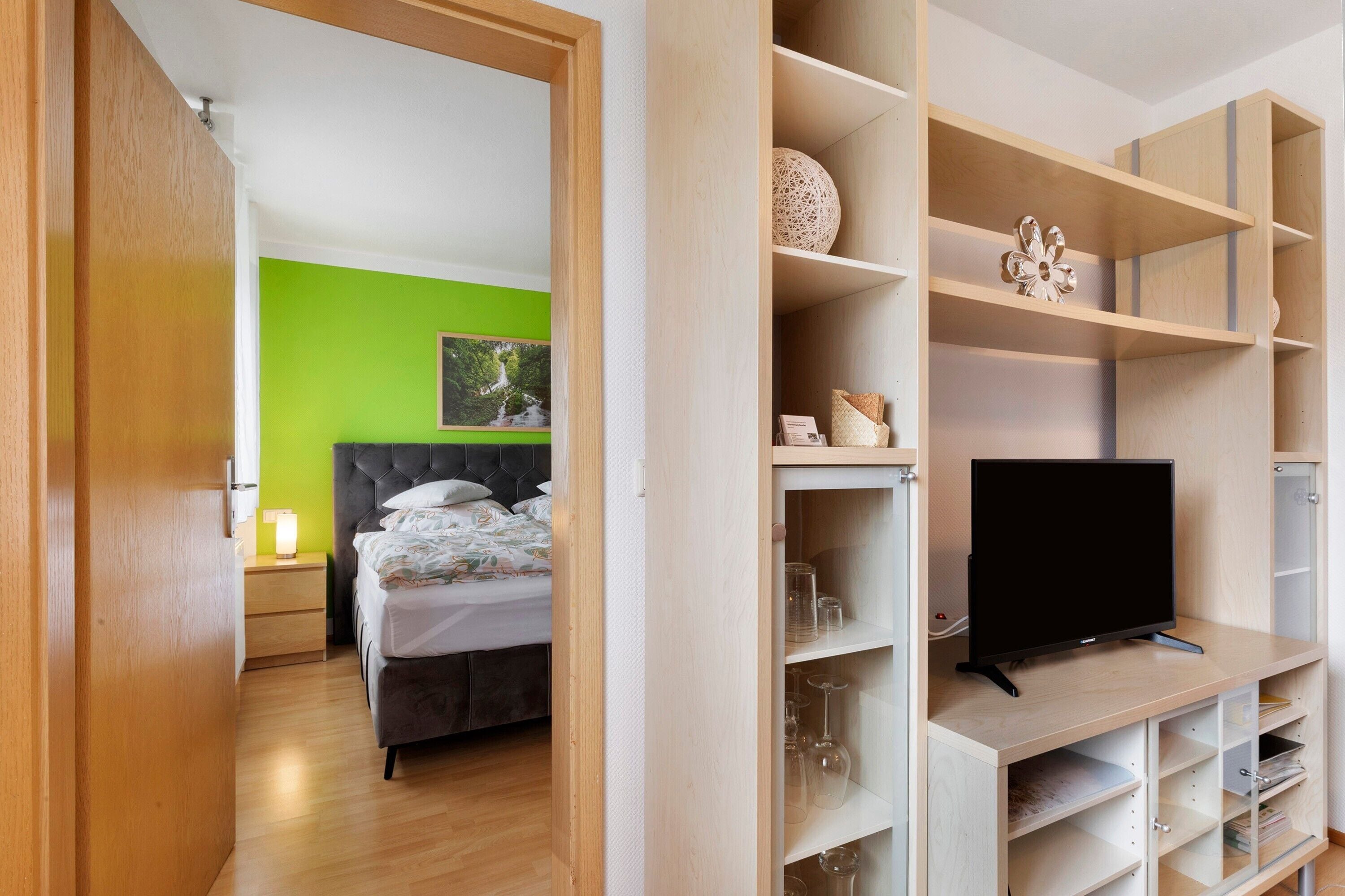A bedroom with a bed, a TV, and a shelf with glasses and decorative items.