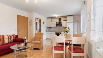 A modern living room with a sofa, chairs, a glass table, and a kitchen in the background.