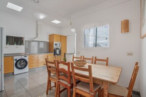 A modern kitchen with a washing machine, wooden cabinets, a dining table with chairs, and a window with a view.