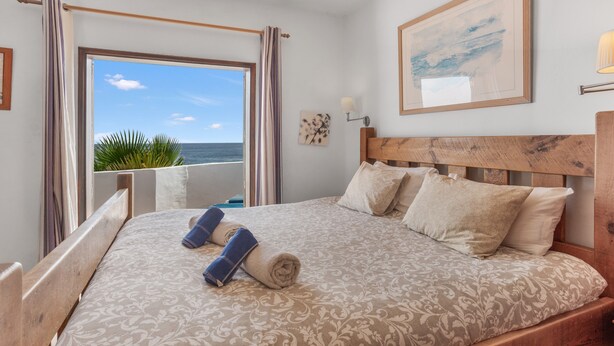 Un dormitorio con una cama grande, una ventana con cortinas, una obra de arte enmarcada en la pared y vista al mar.