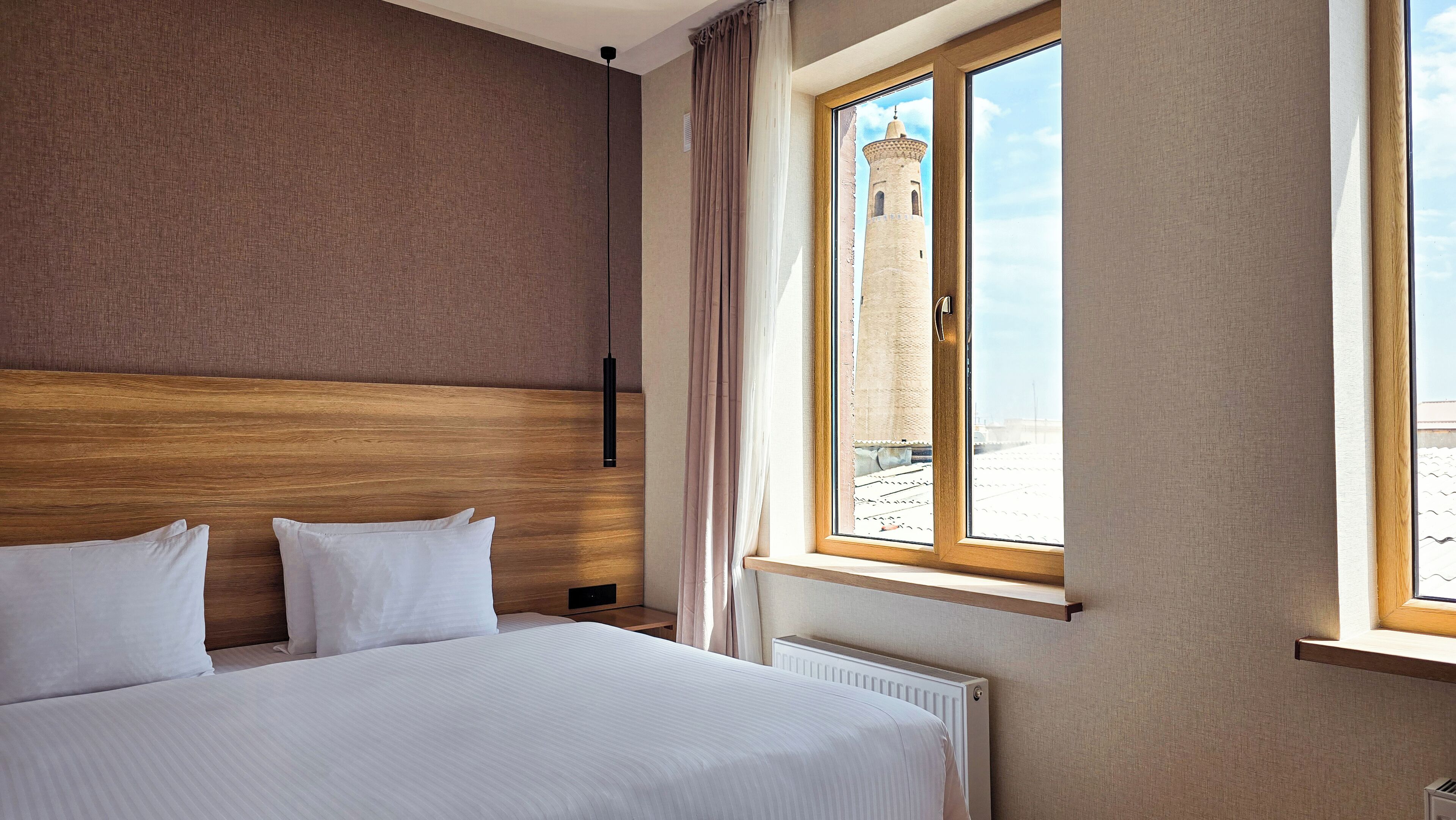 A hotel room with a large bed, a wooden headboard, a window with a view of a tower, and a radiator.