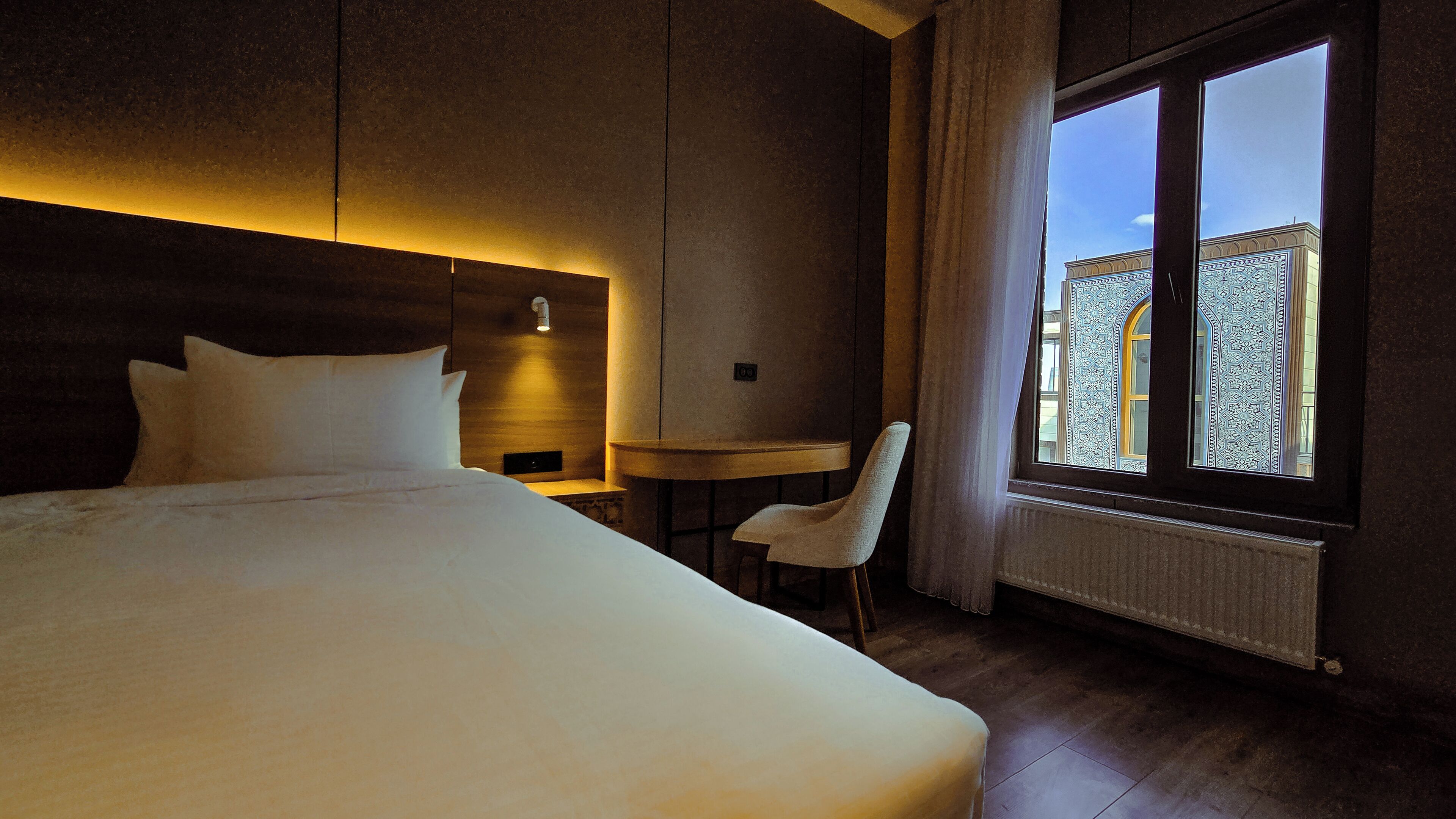 A hotel room with a large bed, a desk, a chair, and a window with a view of a building.
