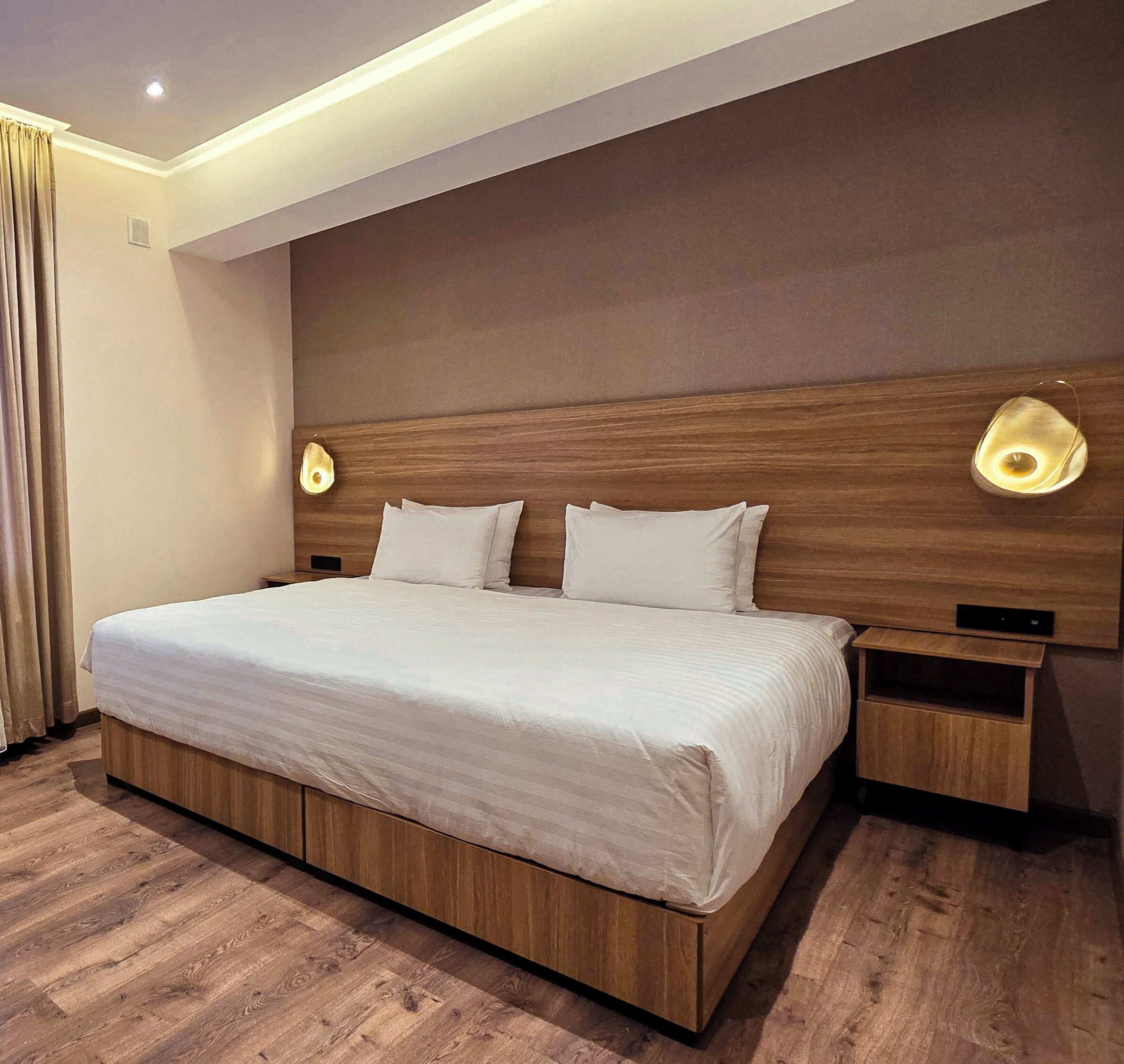 A modern hotel room with a large bed, wooden headboard, two bedside tables with lamps, and a wooden floor.