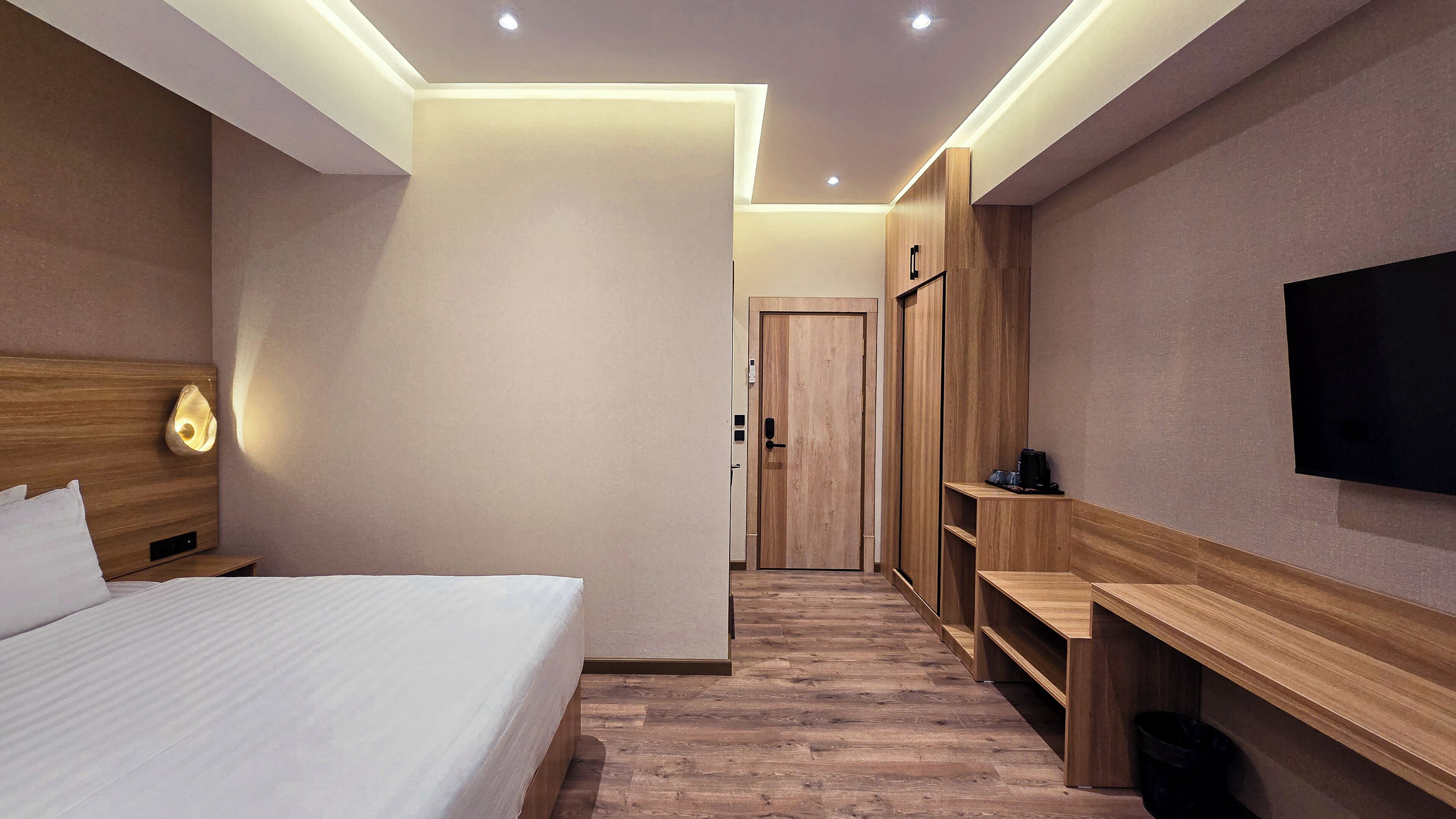A modern hotel room with a large bed, a wooden headboard, a bedside lamp, a flat-screen TV mounted on the wall, and a wooden bench with storage underneath.
