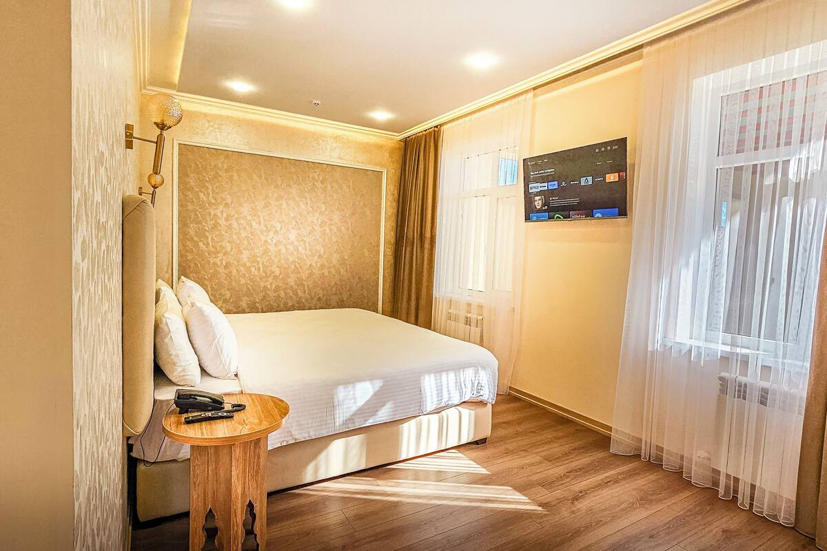 Ein Hotelzimmer mit einem Bett, einem Nachttisch mit Telefon, einem an der Wand montierten Fernseher und transparenten Vorhängen.