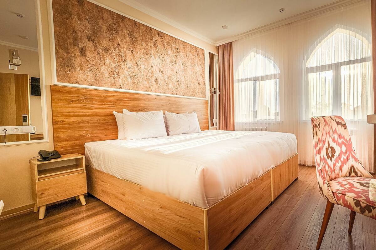 Ein Hotelzimmer mit einem hölzernen Bett, einem Nachttisch mit Telefon, einem Sessel und großen Fenstern mit Vorhängen.