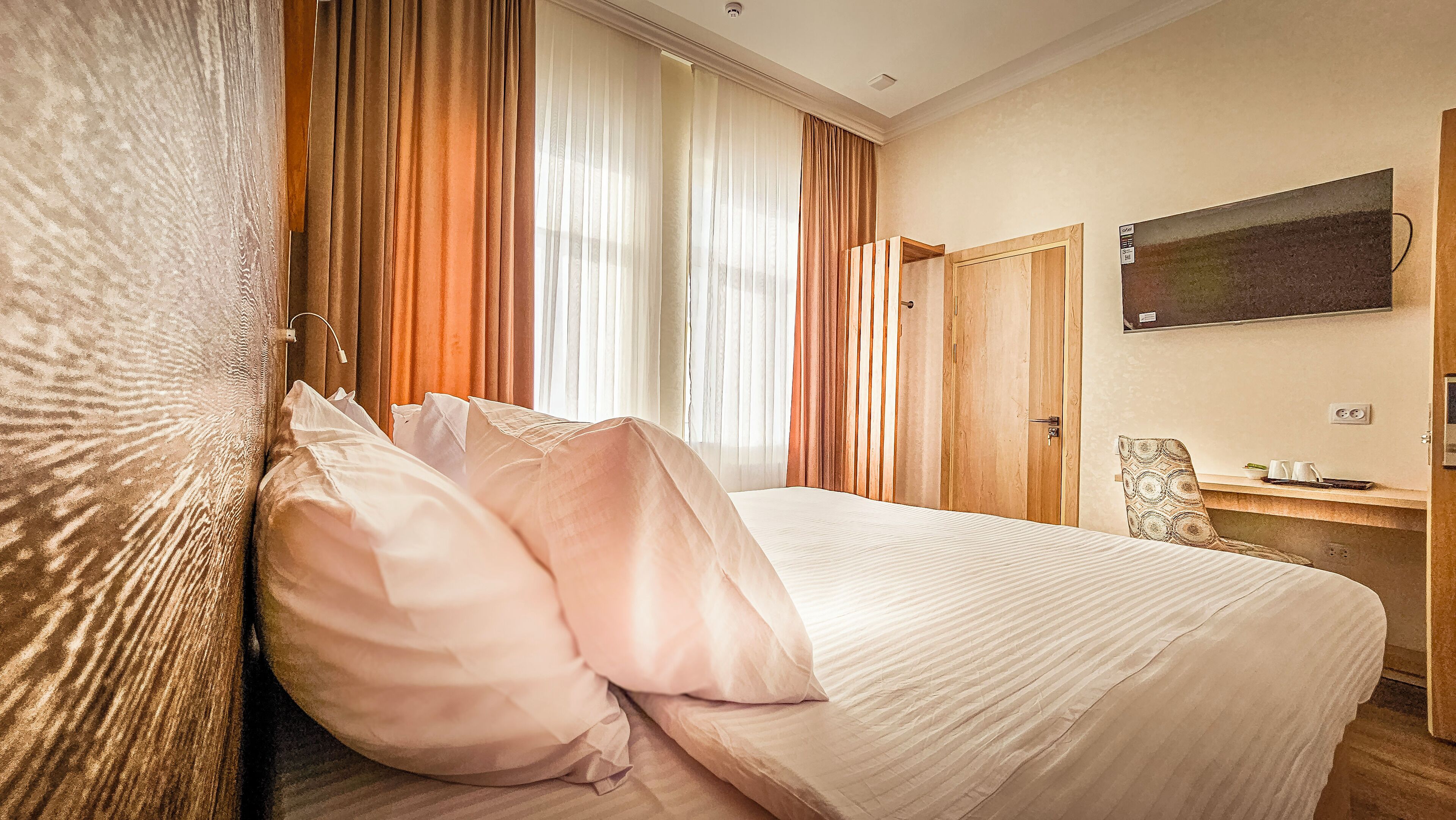 A hotel room with a bed, curtains, a wooden wardrobe, a desk with a chair, and a mounted TV.