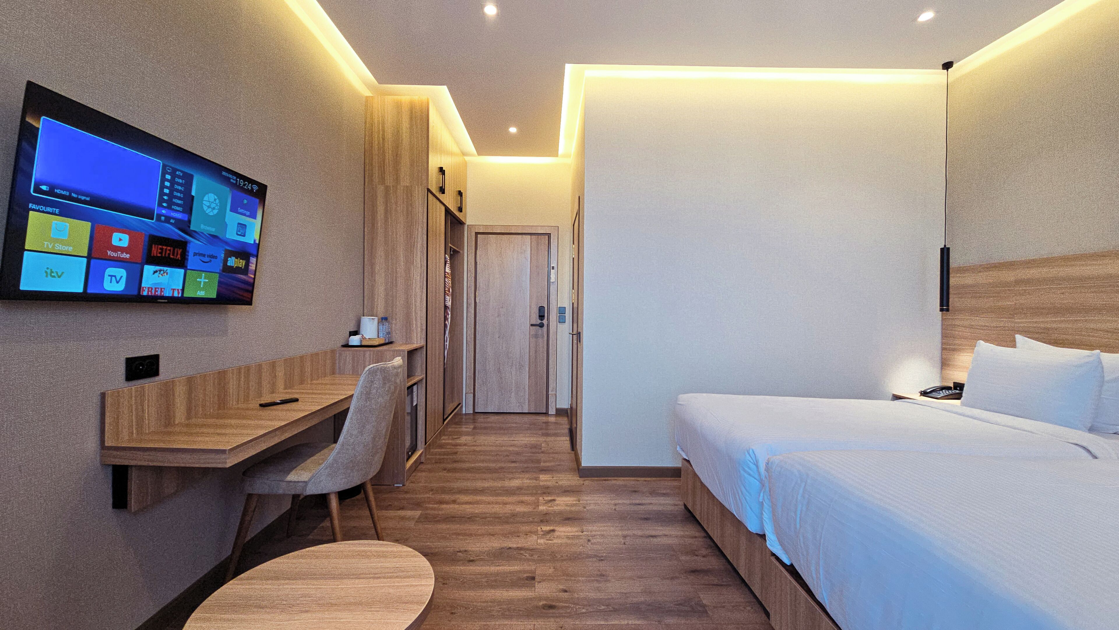 Une chambre d’hôtel moderne avec un bureau en bois, un téléviseur fixé au mur, un lit avec du linge de maison blanc et un plancher en bois.
