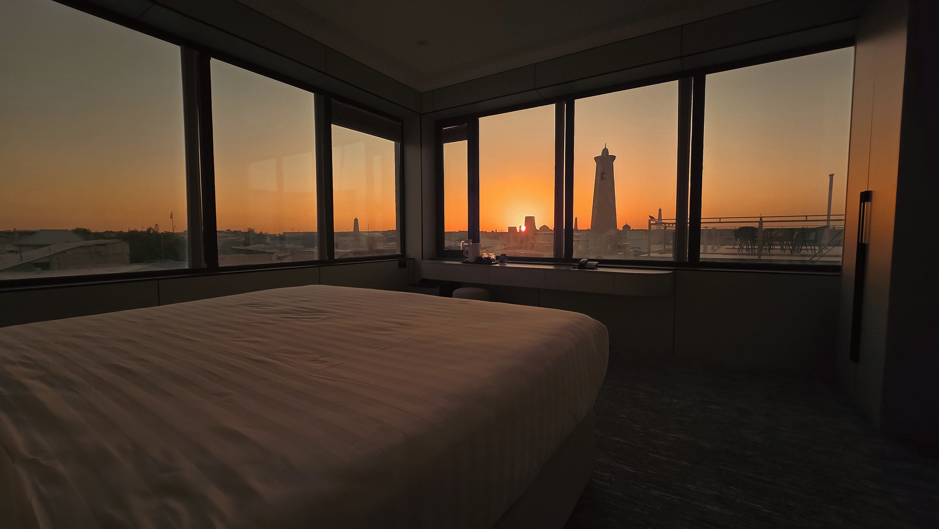 Habitación de hotel con una cama grande, un escritorio y vistas a la silueta urbana al atardecer.